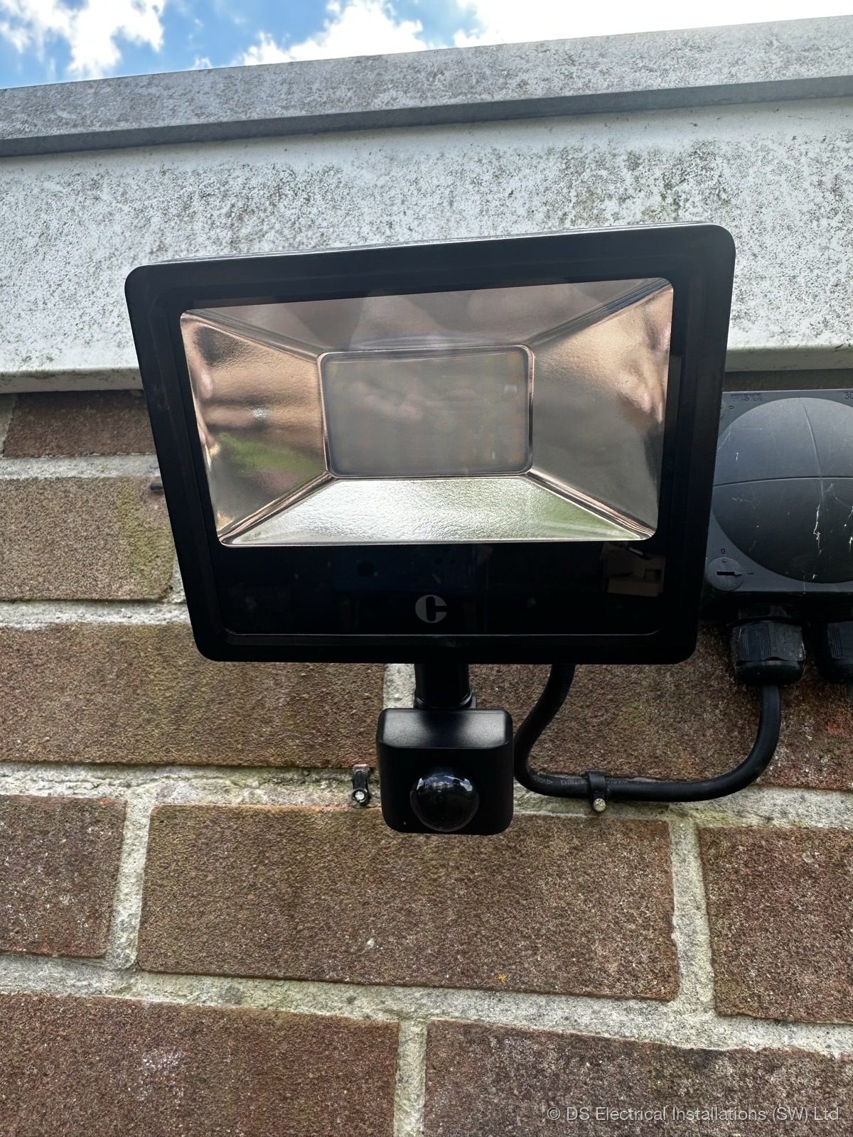 PIR-activated LED floodlight on red brick wall, dusk shot showing beam coverage